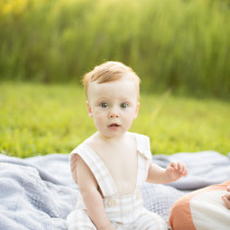 Baby in a gingham romper sitting on a blanket in the grass at sunset