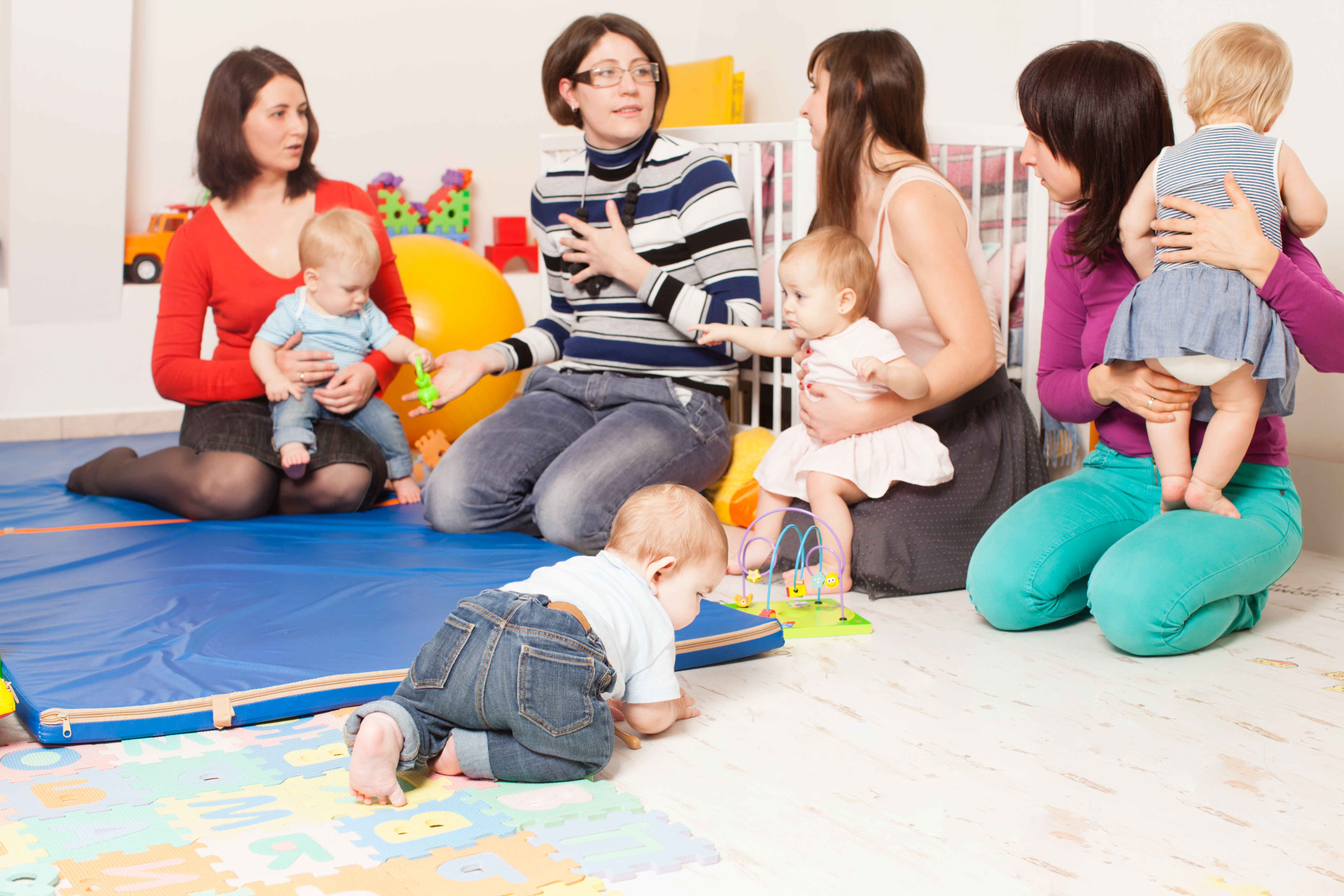 Four moms sitting on a playroom floor with their babies, one baby crawling on a mat