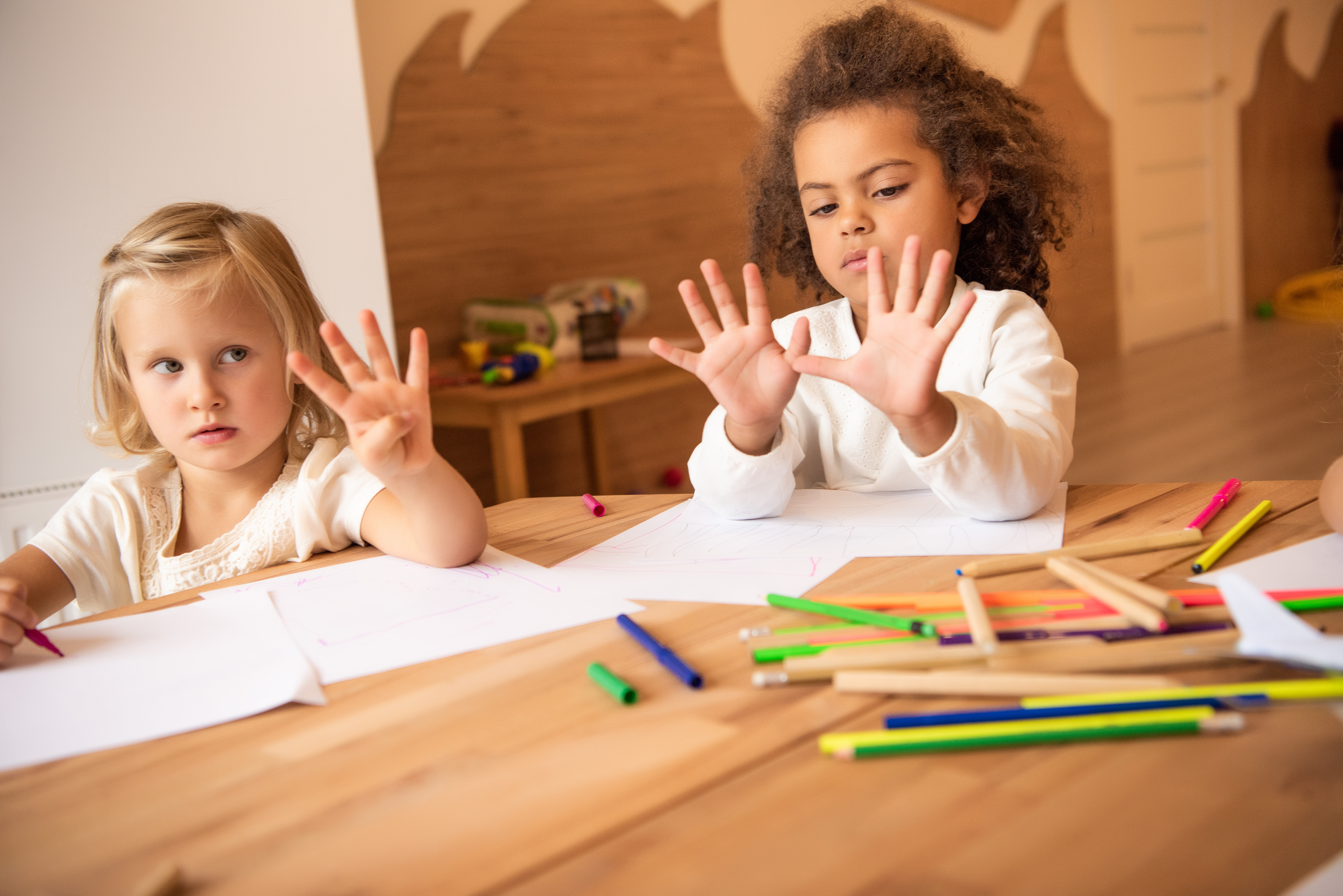 Two preschoolers at a table of markers, holding up their hands with fingers spread