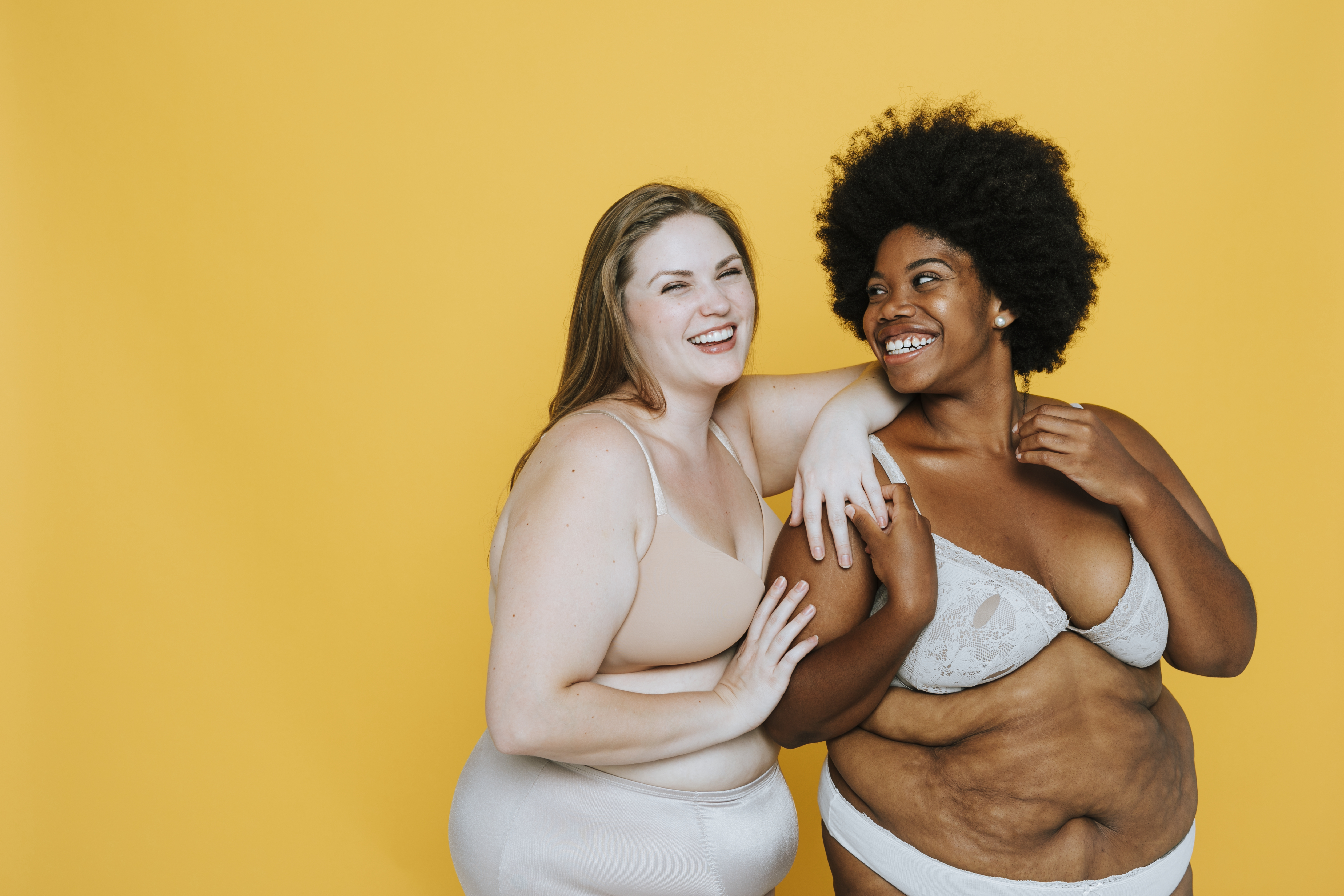 Two laughing women in bras and underwear embracing in front of a yellow backdrop