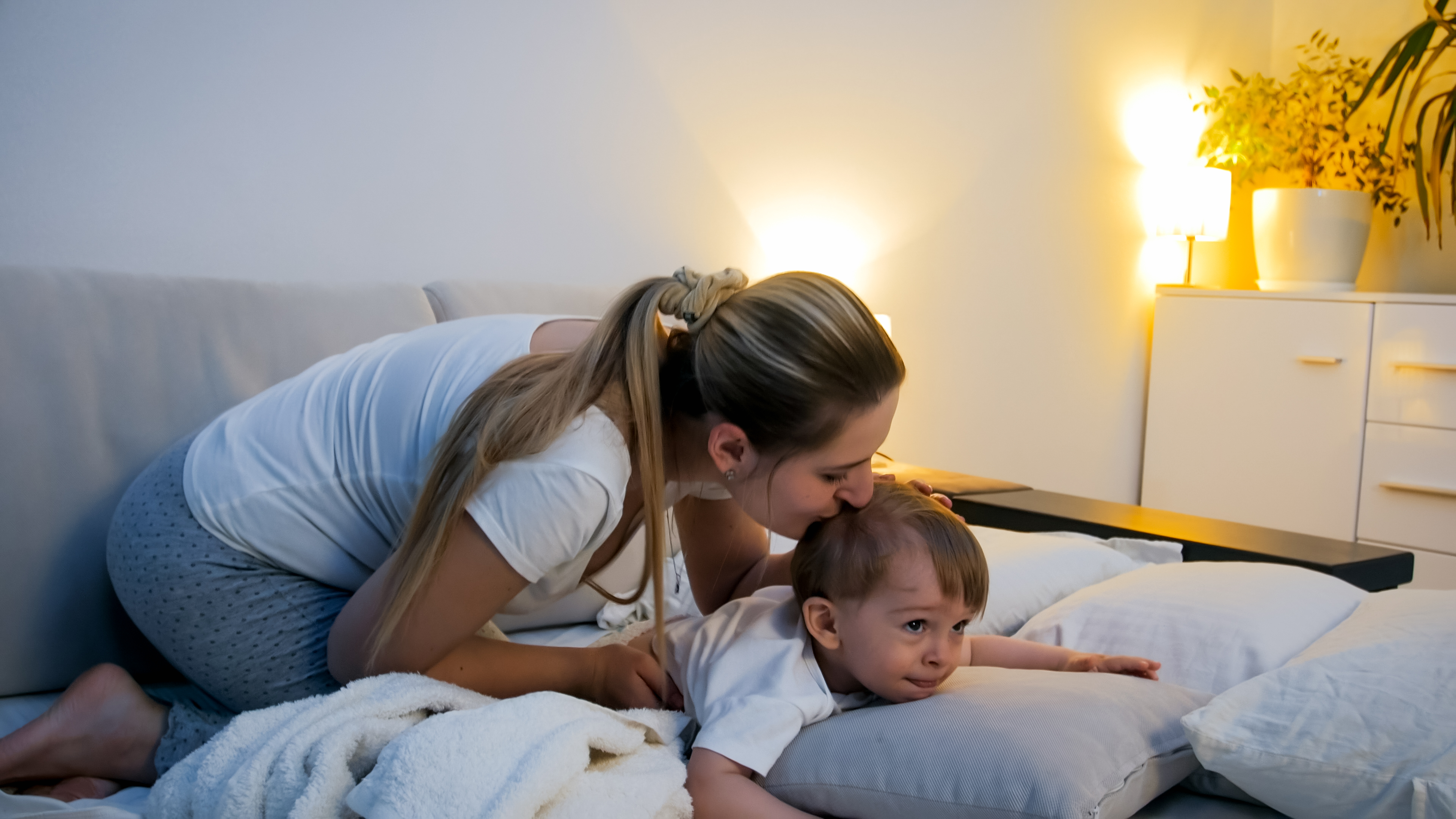 Mom kissing her toddler's head as the child settles onto pillows at bedtime