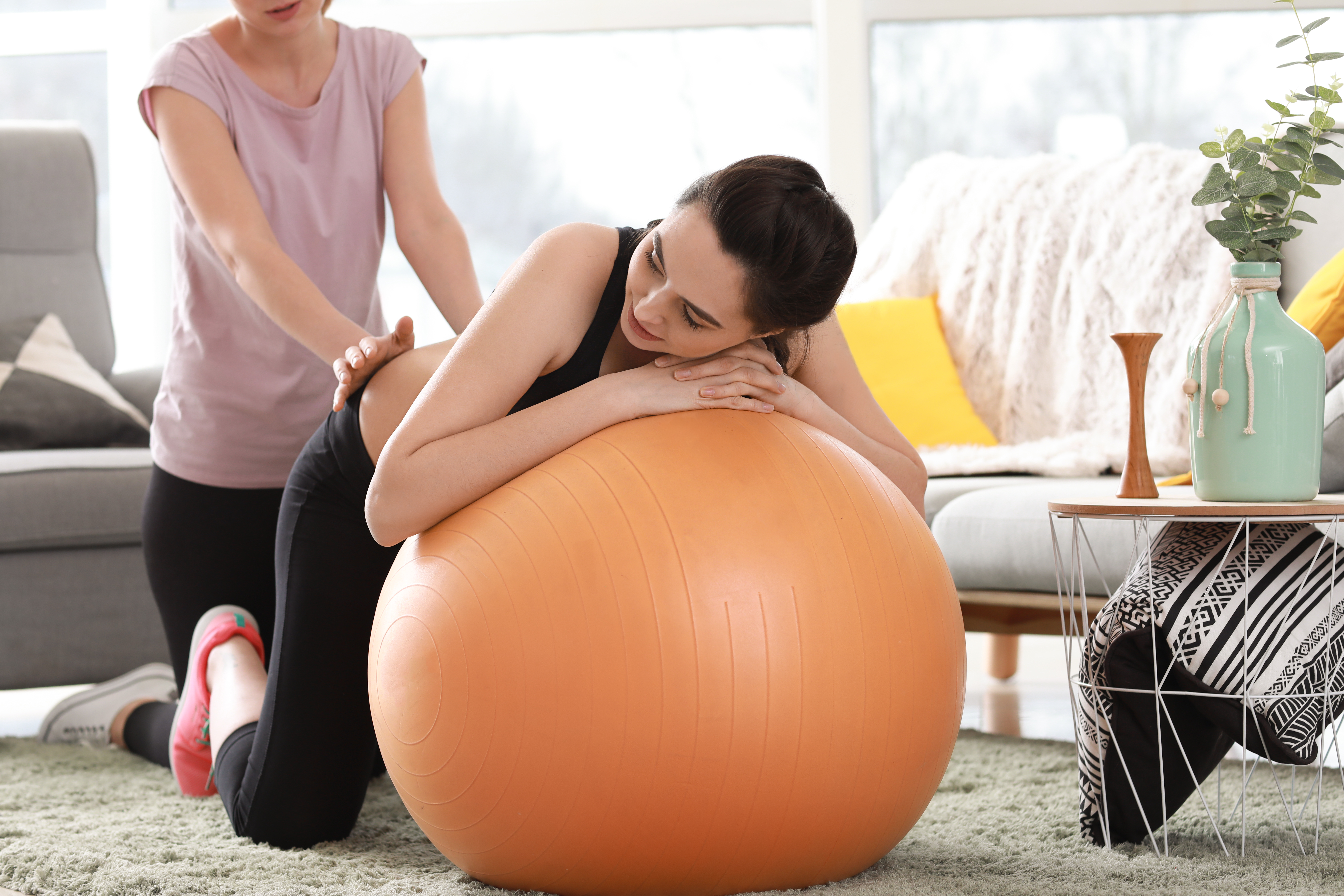 Doula massaging the back of a pregnant woman leaning on an exercise ball at home