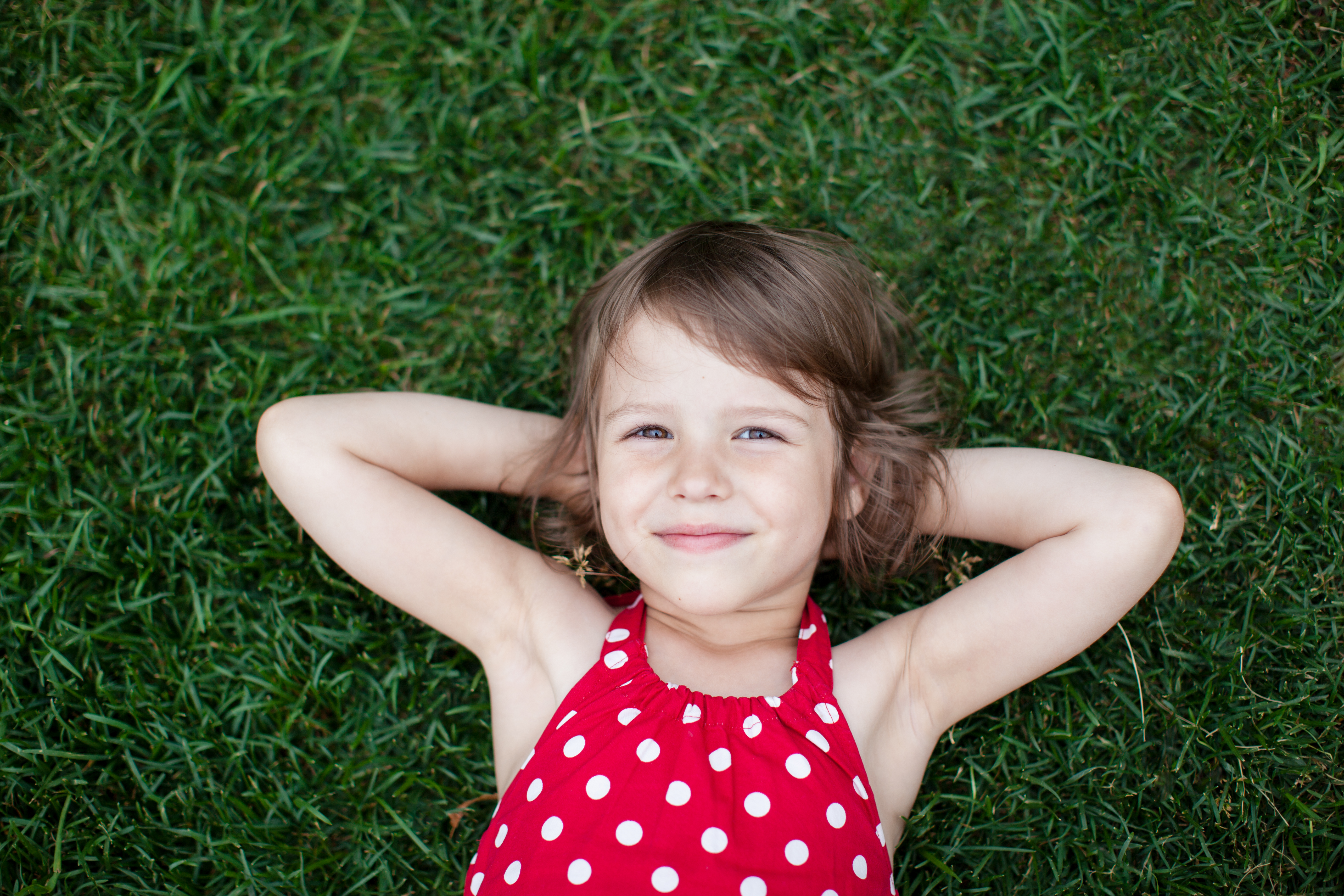Smiling girl in a red polka-dot top lying on the grass with hands behind her head
