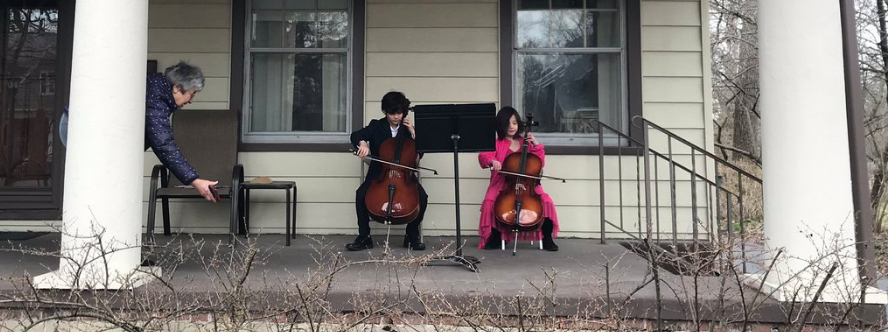 Two children playing cellos on a porch as their elderly neighbor watches from her doorway