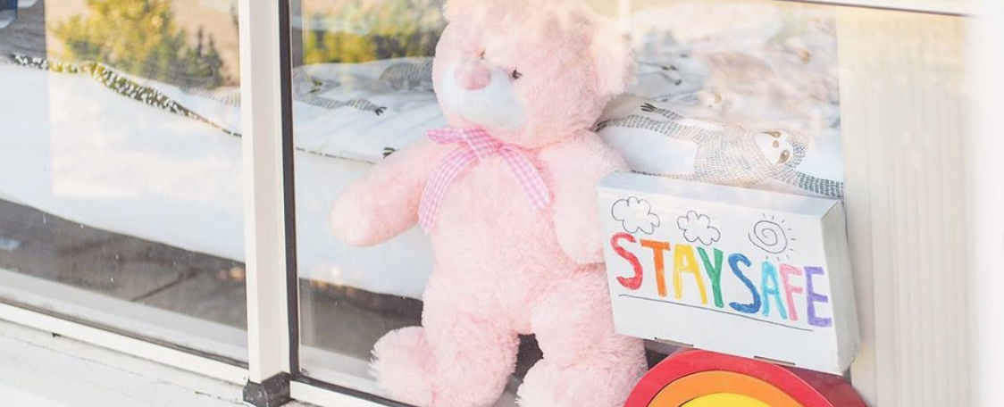 Pink teddy bear in a window beside a hand-drawn rainbow "Stay Safe" sign