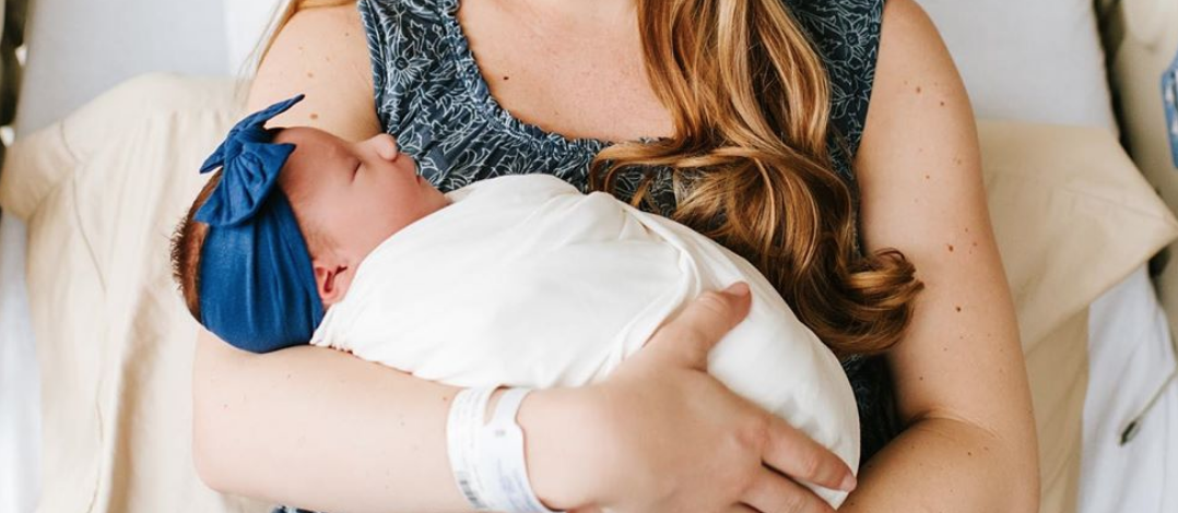 Mom in a hospital bed holding her swaddled newborn, who wears a blue bow headband