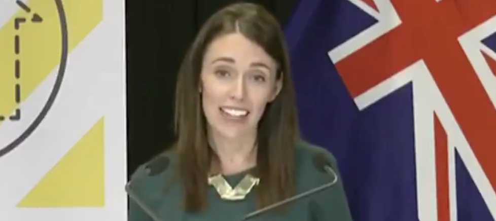 Woman speaking at a press conference podium in front of the New Zealand flag