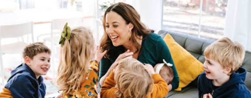 Laughing mom on a sofa holding a baby, surrounded by four young children
