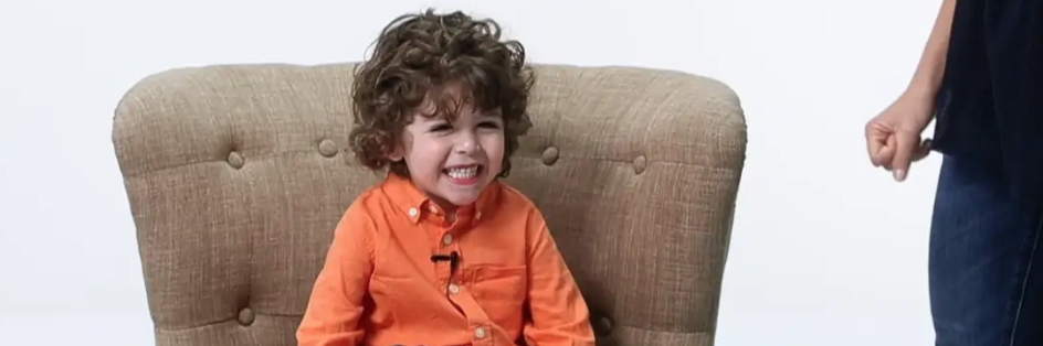 Grinning curly-haired little boy in an orange shirt sitting in an armchair, from the Project M video
