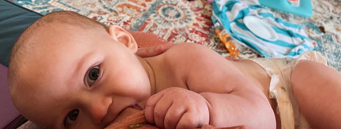 Baby lying on a rug chewing on a cloth, looking up at the camera