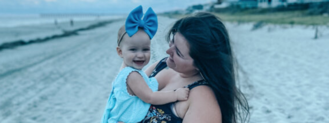 Mom on the beach holding her smiling baby, who wears a big blue hair bow