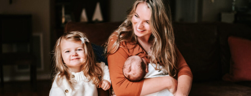 Smiling mom holding her newborn on a couch next to her young daughter