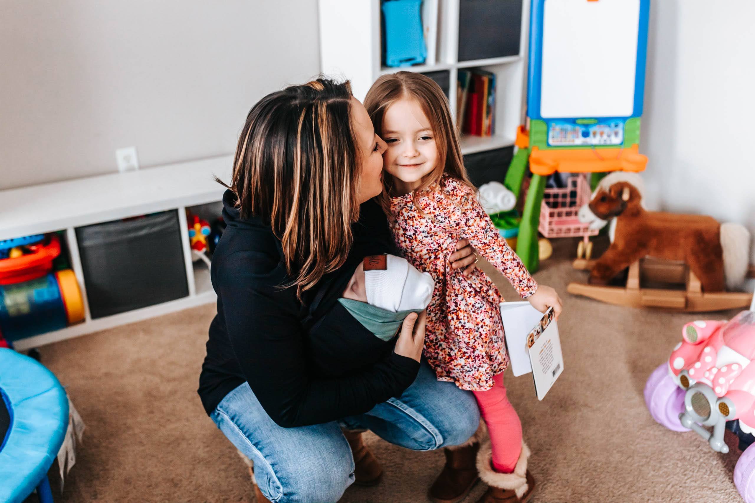 Mom kneeling in a playroom holding a newborn and kissing her young daughter on the cheek