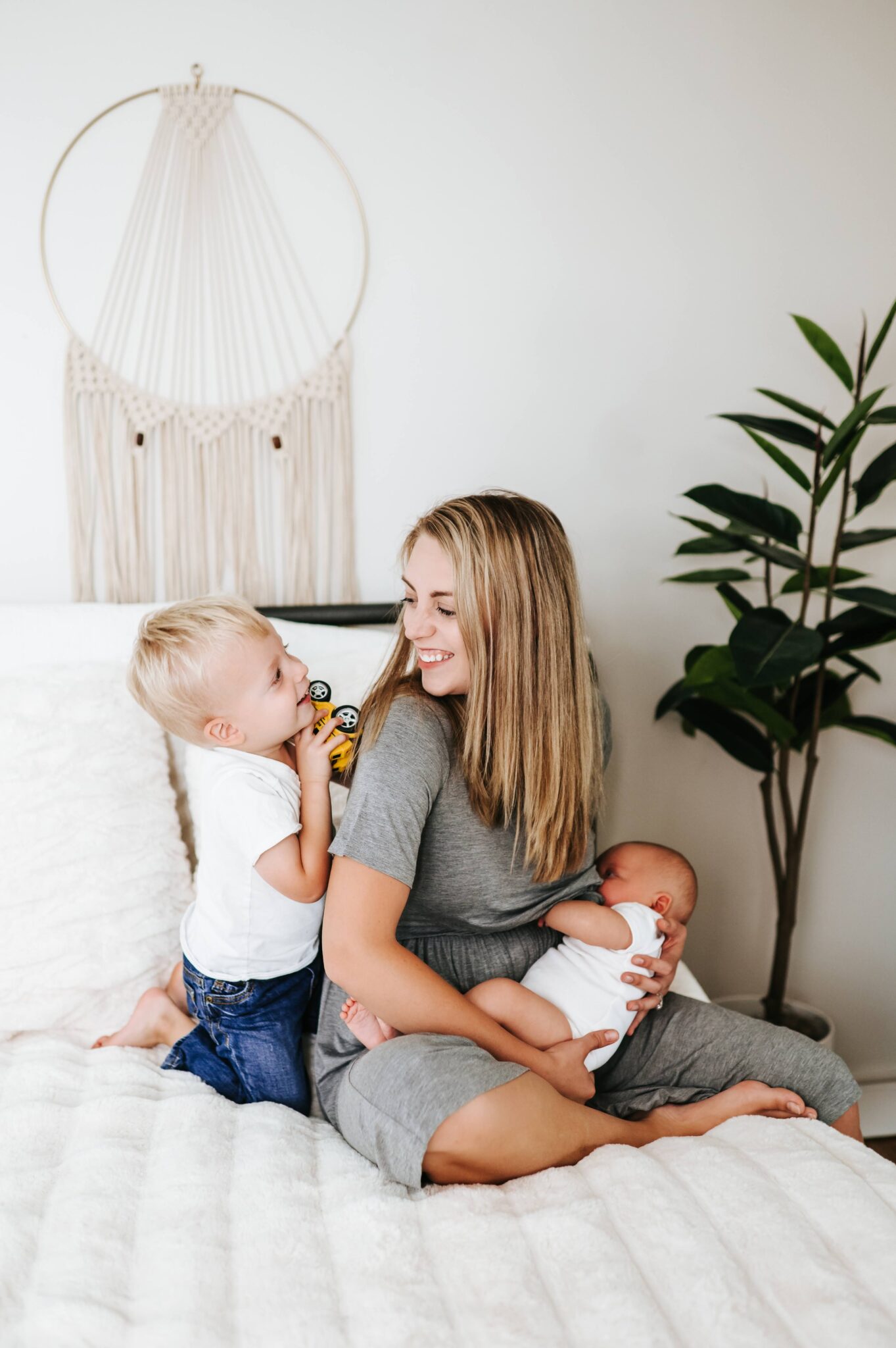 Smiling mom sitting on a bed holding her newborn while her toddler son plays with a toy truck beside her