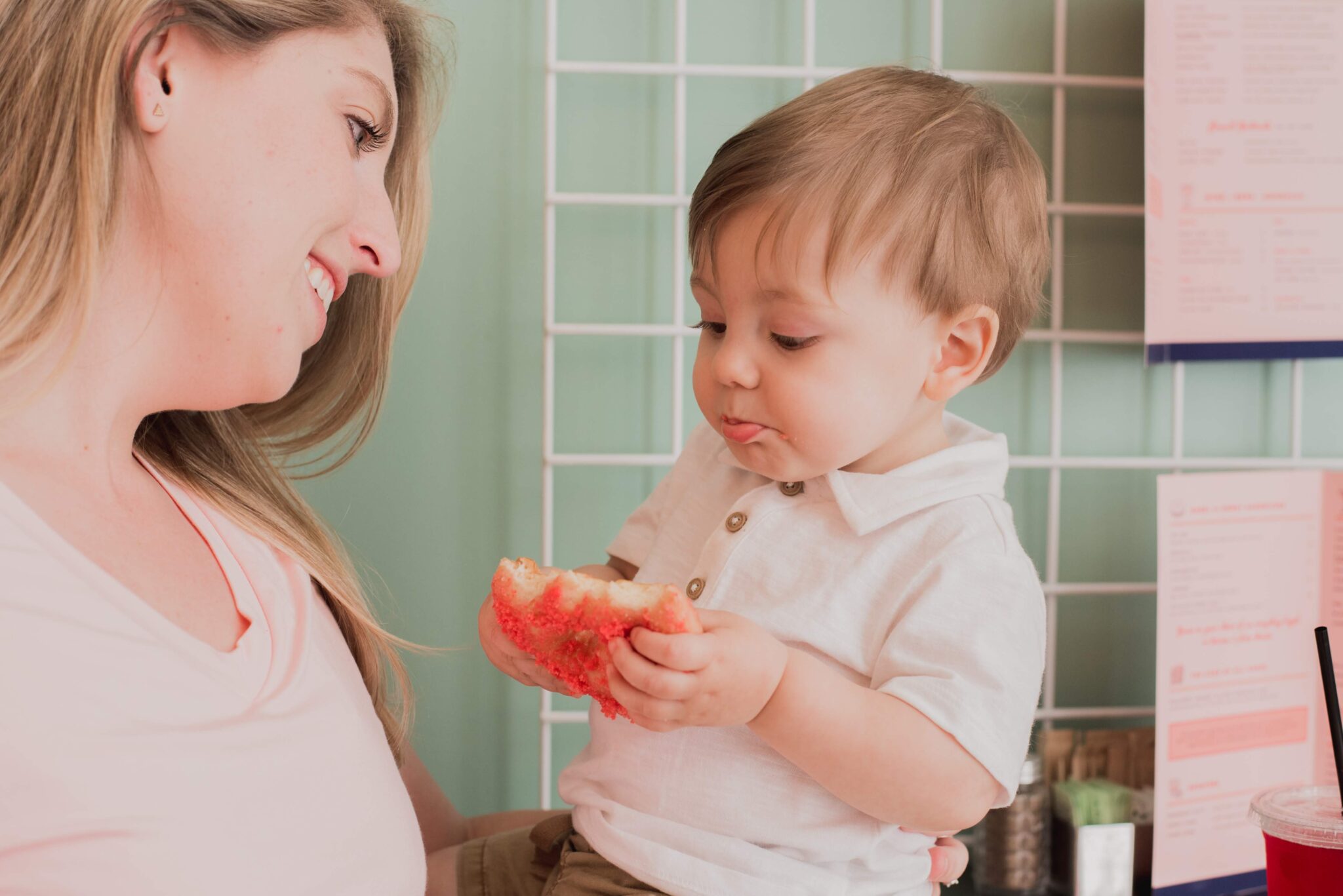 Smiling mom watching her baby boy hold and taste a pink, sugary pastry